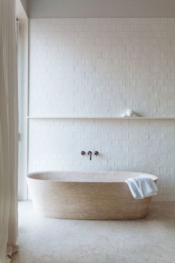 a neutral bathroom with white tiles, a stone bathtub and a floating shelf for an airy and serene feeling in the space