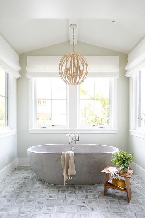 a neutral farmhouse bathroom with printed tiles, a neutral stone bathtub and a round chandelier looks serene