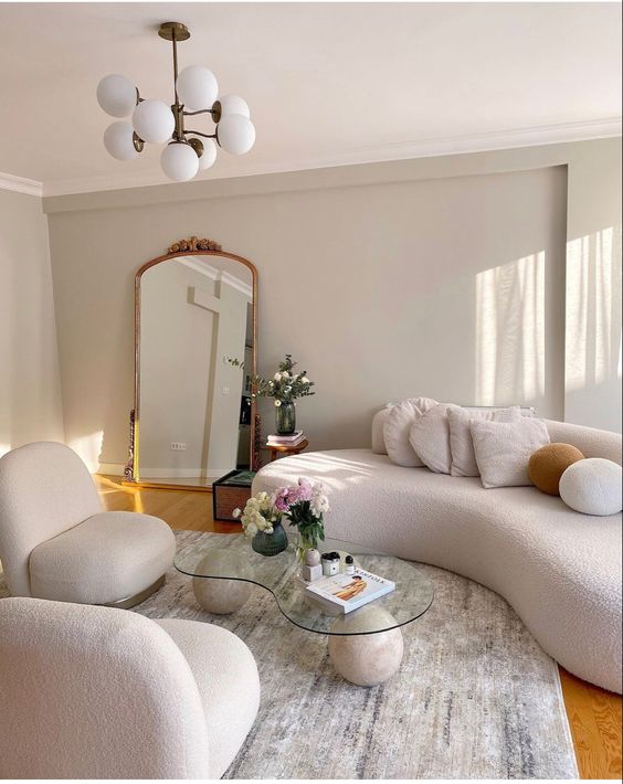 a pretty neutral living room with creamy boucle curved furniture, a matching glass coffee table, a mirror in a gold frame