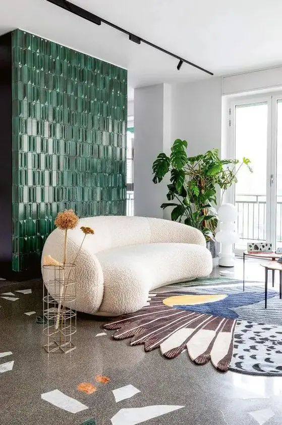 a quirky living room with a green tile wall, a white boucle curved sofa, a colorful rug, side tables and greenery