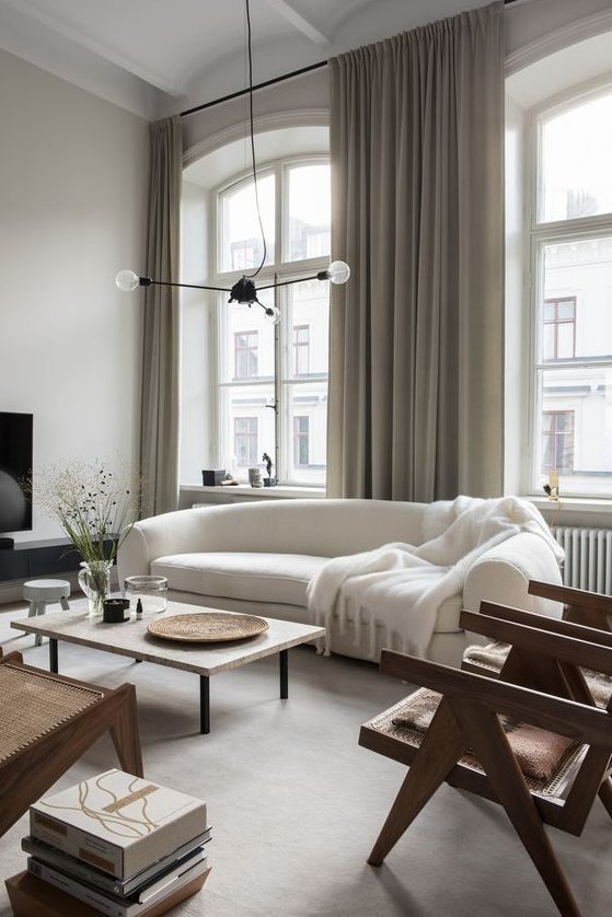a refined Scandinavian living room with a curved white sofa, rattan chairs, a low coffee table, a pendant lamp and books