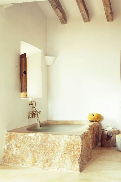 a rustic bathroom with white walls, wooden beams, a rough stone bathtub in a warm shade and tree stumps as side tables