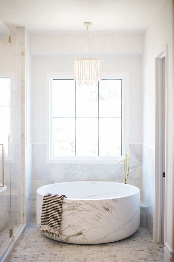 a small yet refined neutral bathroom clad with marble tiles, with a marble half wall and a marble round bathtub