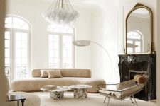 a sophisticated neutral living room with a black marble fireplace, a neutral curved sofa, a chair, marble coffee tables and a bubble chandelier