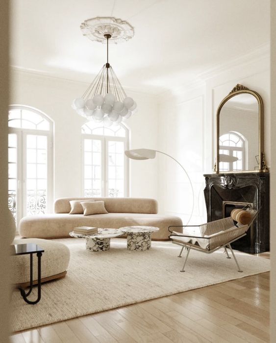 a sophisticated neutral living room with a black marble fireplace, a neutral curved sofa, a chair, marble coffee tables and a bubble chandelier