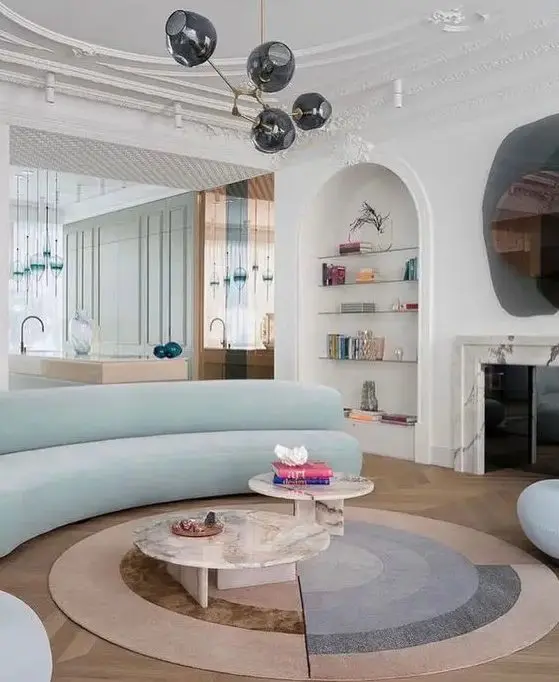 A whimsical living room with a white marble fireplace, built in shelves, a light blue curved sofa, marble tables and cool rugs