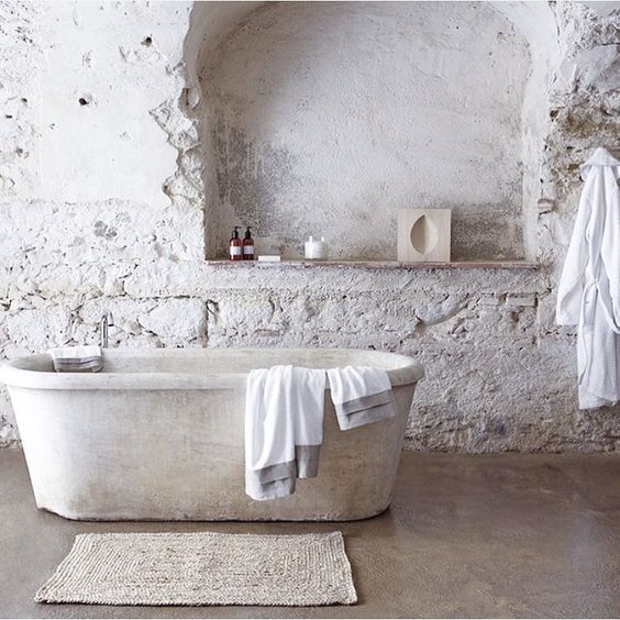 A white bathroom with rough stone tiles, a white stone bathtub and a jute rug is all natural and relaxing