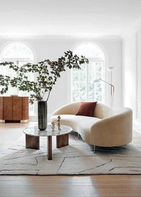 an airy Scandinavian living room with arched windows, a creamy curved sofa, a chic credenza and a three-leg round table, a floor lamp