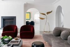 an exquisite living room with a fireplace, a grey curved sofa with pillows, burgundy curved chairs and rounded tables