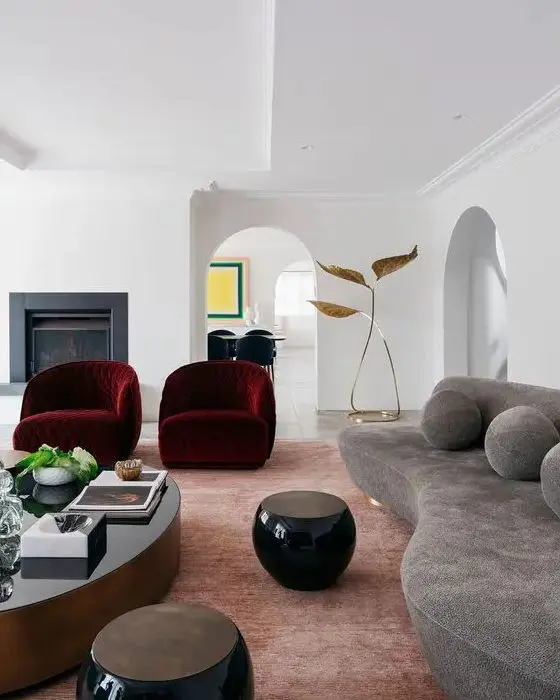 an exquisite living room with a fireplace, a grey curved sofa with pillows, burgundy curved chairs and rounded tables