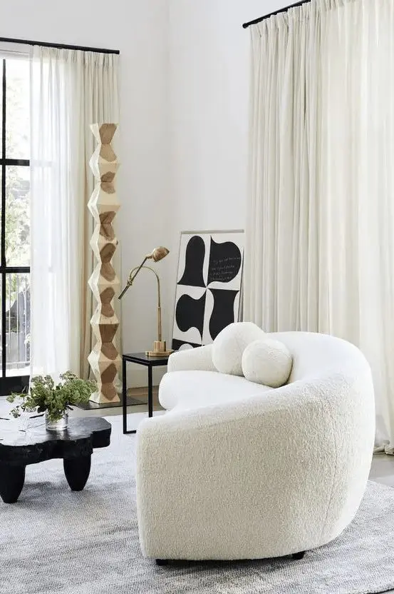 An eye catchy space with a white boucle curved sofa, a black tree slice table, a side table with a lamp and some abstract art