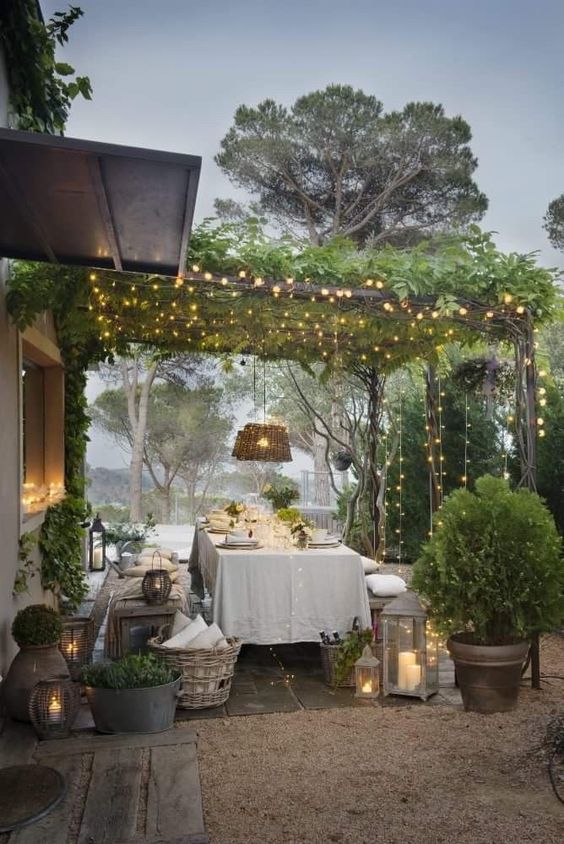 an outdoor dining room with a canopy of greenery and lights, candle lanterns, lots of potted greenery and wicker touches