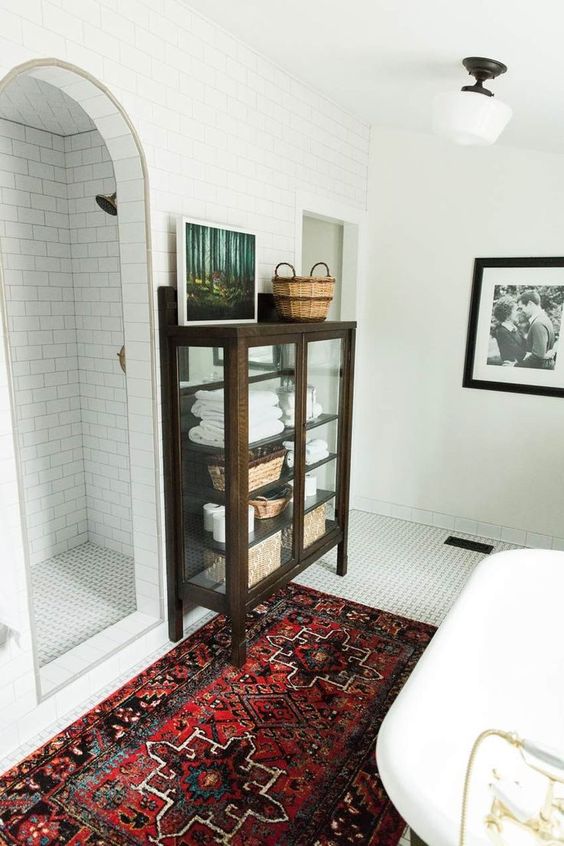 a modern bathroom with a bright rug, white tiles all over and a vintage dark stained glass armoire