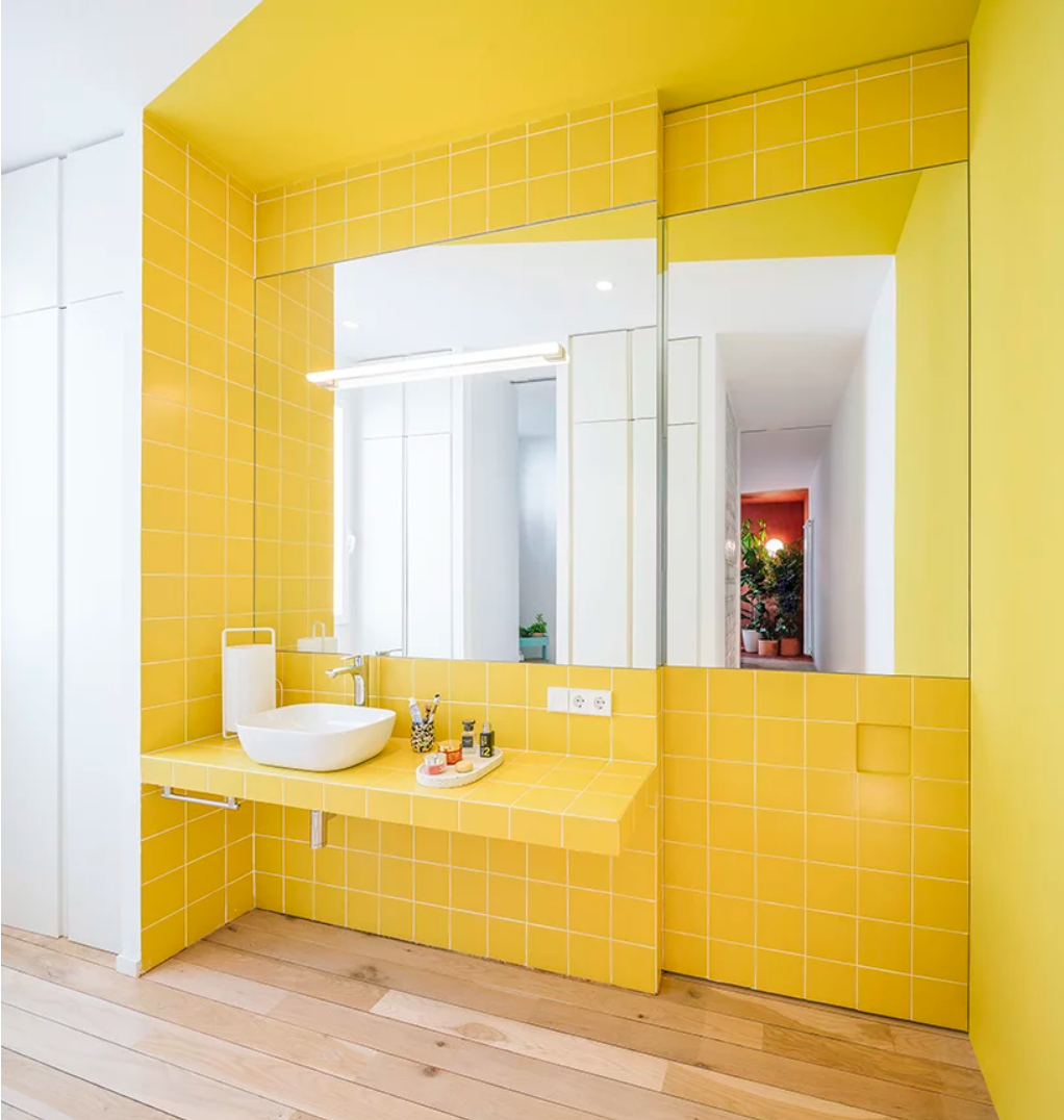 The color block bathroom is done with bright yellow tiles that also form a vanity and there are two large mirrors