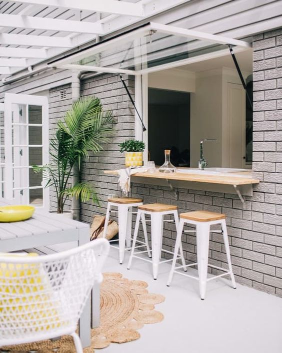 An outdoor indoor bar with stools, a countertop and a dining space by its side plus some potted greenery
