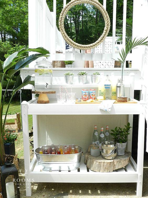 a white home bar station with various drinks, a cooler of a bucket and potted succulents and greenery