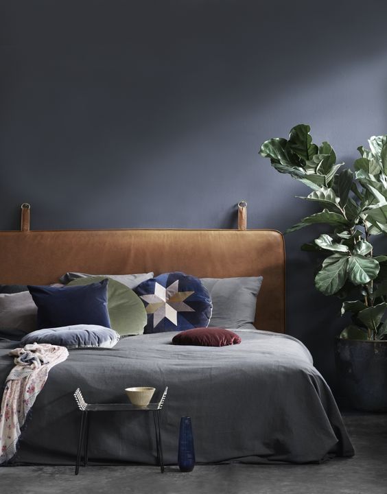 a moody bedroom in black refreshed with a potted plant, bright pillows and a leather hanging headboard for a statement