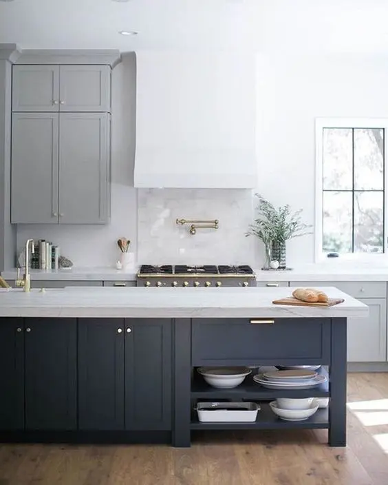 a beautiful and airy contemporary kitchen with grey cabinets, a white hood, a navy kitchen island with a stone countertop and gold touches