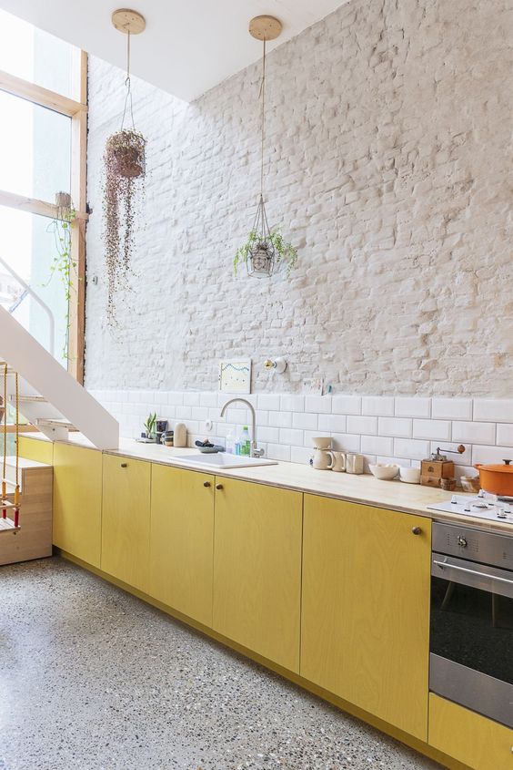 a bold contemporary kitchen with yellow plywood cabinets, neutral countertops, a white subway tile backsplash and a brick wall