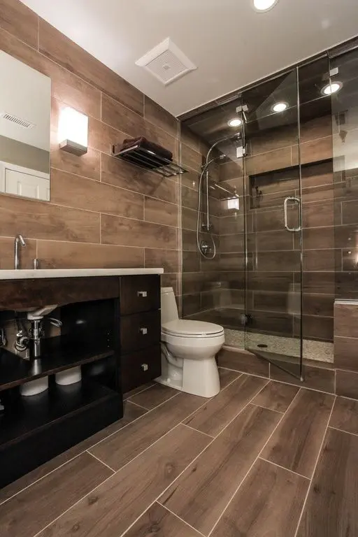 A cozy and cool bathroom completely clad with wood tiles, with a shower space, a dark vanity and a mirror plus built in lights