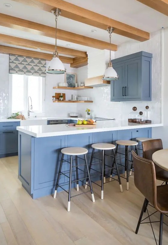 A fab coastal kitchen with blue cabinets, white stone countertops and glossy tiles, light stained shelves and wooden beams