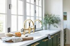 a hunter green modern kitchen with white stone countertops, gold handles and fixtures and a window instead of a backsplash