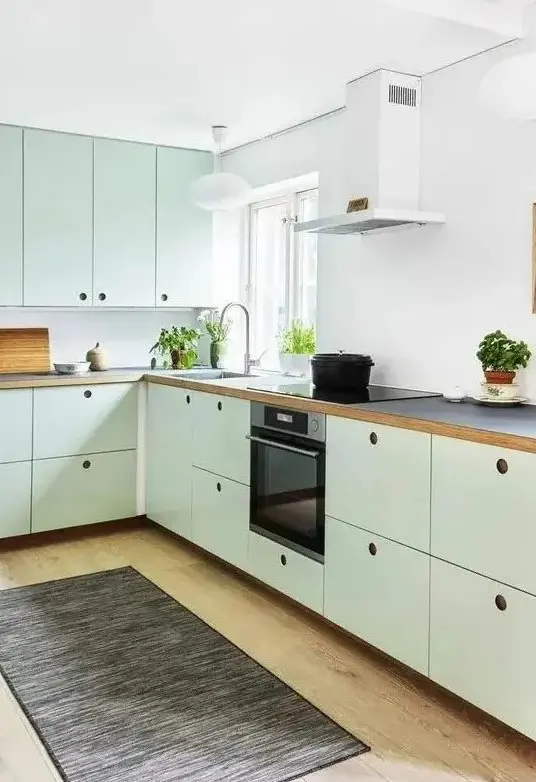 A mint green Scandinavian kitchen with MDF cabinets, black countertops and a white backsplash, a dark rug and built in appliances
