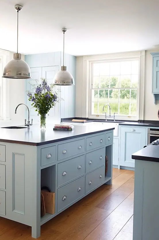 a modern light blue country kitchen with dark countertops, pendant lamps and large windows plus a lovely hardwood floor is chic
