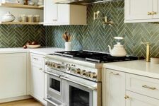 a refined kitchen with creamy cabinets, a green herringbone tile backsplash, a white hood and gold sconces