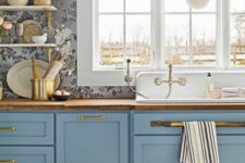 a refined kitchen with grey floral print wallpaper, blue shaker cabinets, butcherblock countertops, gold handles and a gold pendant lamp