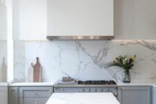 a serene grey and white kitchen with only a lower row of cabinets in grey, a white hood, a white marble backsplash and a matching kitchen island