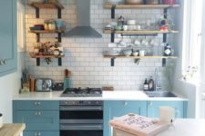 a small blue kitchen with a white tile backsplash and white countertops plus wooden shelves