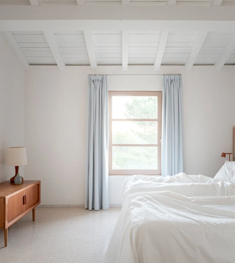 The bedroom is very airy, with a mid-century modern sideboard, a bed, sconces and blue curtains