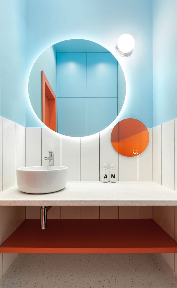 a bold bathroom with light blue half walls, an orange shelf and an orange round mirror for a bright look