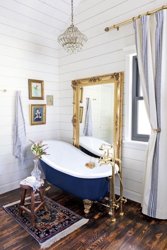 a bathroom covered with white shiplap, with a mirror in an ornate gold frame, artworks, blue tub on gold legs