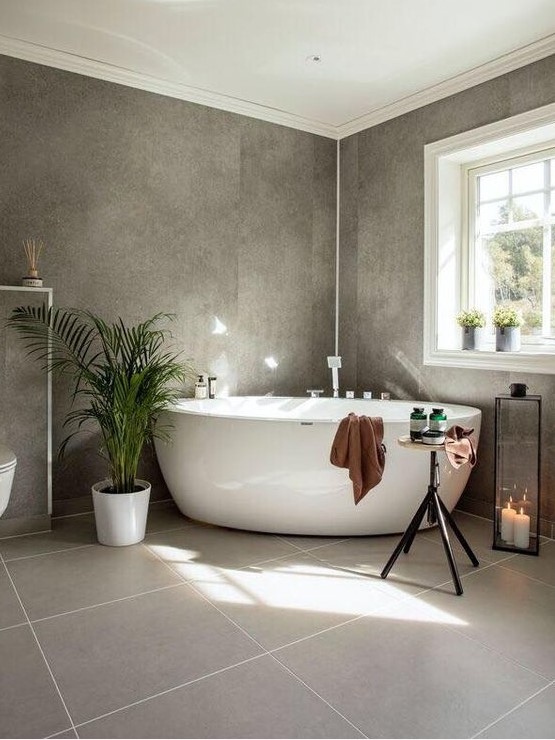 A beautiful grey bathroom with two kinds of tiles, potted plants, a free standing tub, a candle lantern and a table