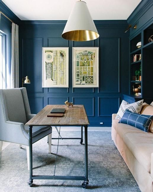 a blue home office with paneling, some comfy furniture, an industrial desk, bold artworks and a catchy white and gold lamp
