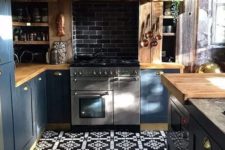 a blue kitchen with light-colored wooden countertops, a wooden hood and a dark tile backsplash plus a vintage lamp