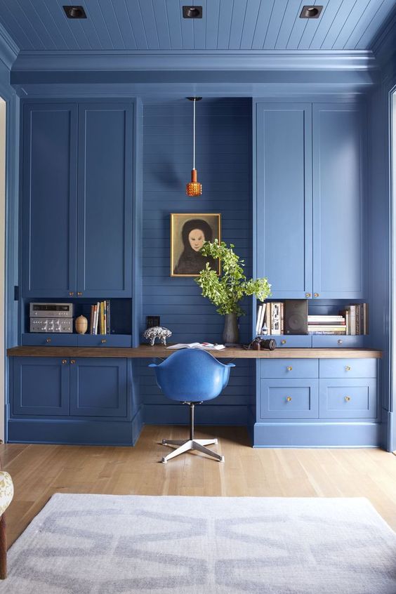 A catchy blue home office with large storage units, a built in desk, a pendant lamp and greenery in a vase