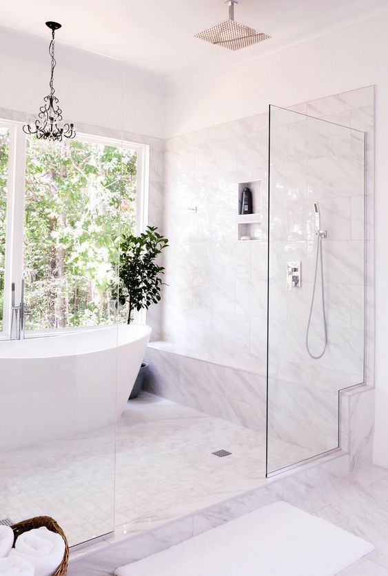 a chic and glam white bathroom clad with marble tiles, a large shower space, a tub, a chandelier and a potted plant