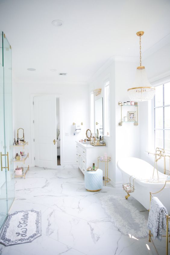 a chic and glam white bathroom with a shower space, a tub by the window, a crystal chandelier, gold fixtures and some decor
