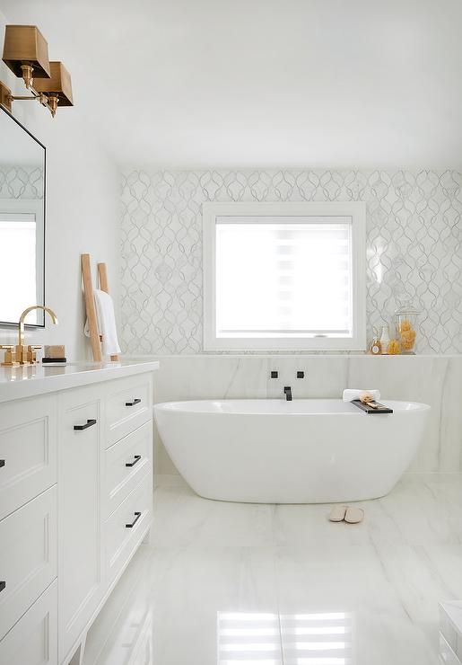 a chic and refined white bathroom done with white marble, a tub, a vanity, a mirror and brass fixtures