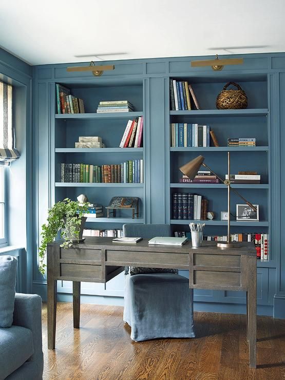 A chic blue home office with built in storage units, a weathered wooden desk, gold and brass touches