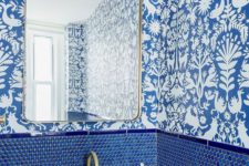 a chic powder room with blue and white wallpaper, blue penny tiles, gold fixtures and lamps plus a stone sink