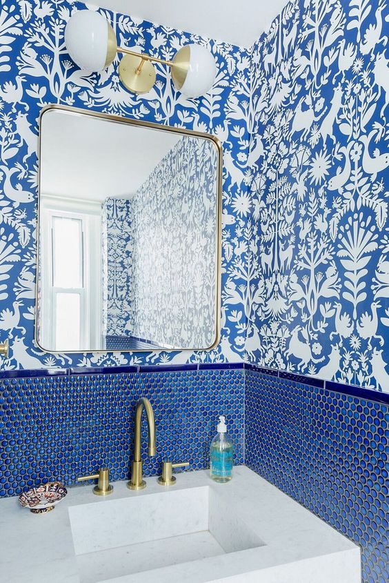 a chic powder room with blue and white wallpaper, blue penny tiles, gold fixtures and lamps plus a stone sink