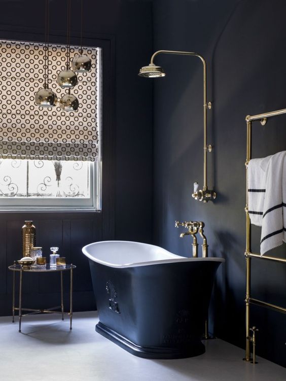 a chic vintage bathroom with black paneled walls, a black tub, gold fixtures, printed shades and gold accessories
