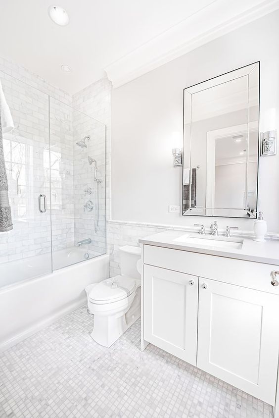 a chic white bathroom done with marble tiles, a tub shower space, a vanity, a mirror and stainless steel fixtures