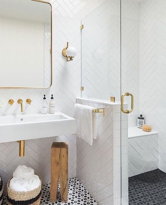 a chic white bathroom with white tiles clad in a herringbone pattern, gold handles, fixtures, frames and sconces