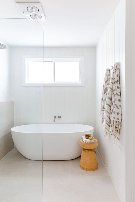 a clean coastal bathroom with a tub by the window, a cork stool and a shower space, printed textiles
