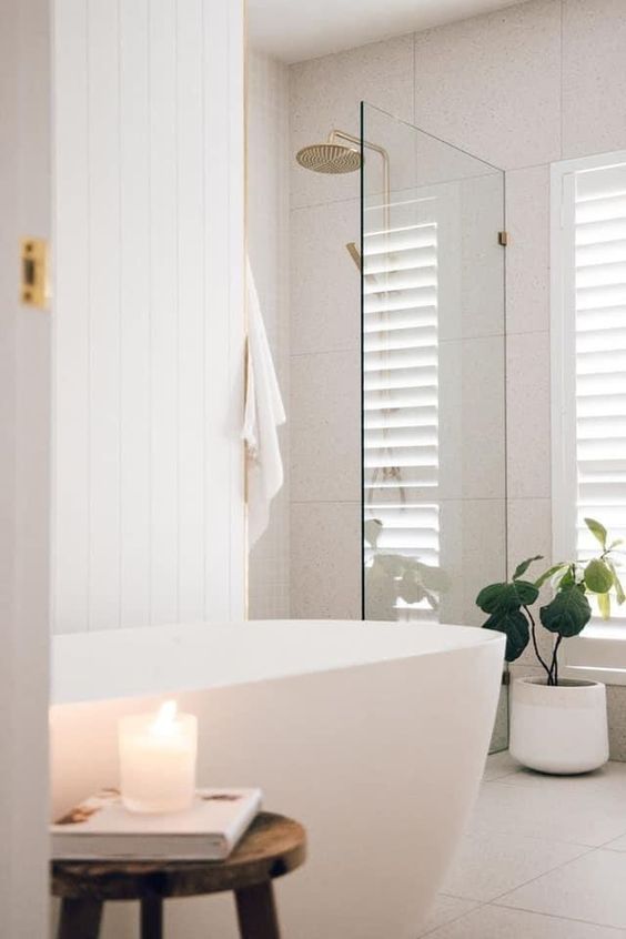 a clean white bathroom with planked and tile walls, a shower space, a tub, a stool, a potted plant and gold fixtures